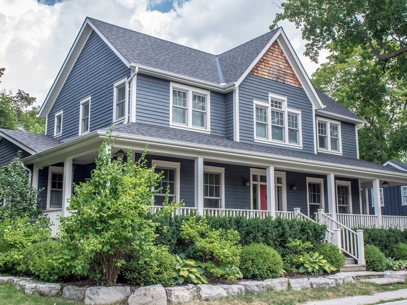 Pictured is a two-story single-family home clad in blue siding with white trim accents with a red front door and lush green landscaping
